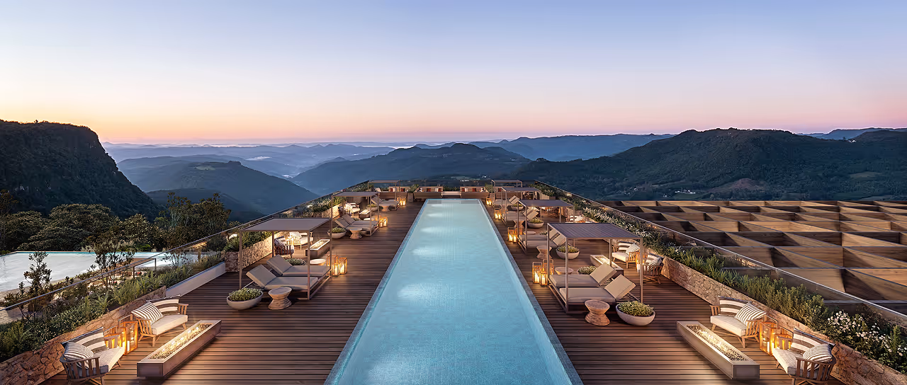 Piscina moderna em deck de madeira com espreguiçadeiras e iluminação, vista para montanhas ao pôr do sol.