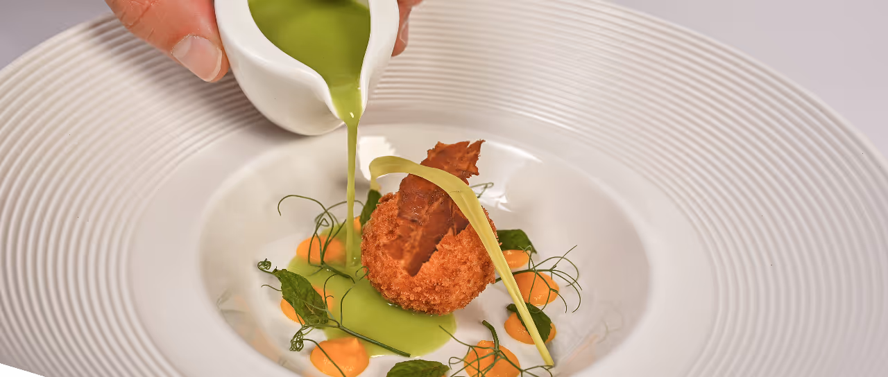 Mão despejando molho verde sobre bolinho frito decorado com ervas e purê laranja em prato branco.