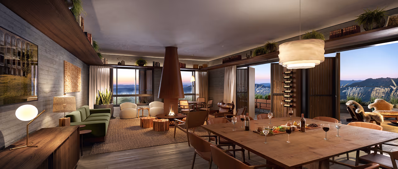 Sala de estar aconchegante com sofá verde, poltronas, lareira central, mesa de jantar com comidas e vinhos, e grandes portas de vidro abertas para uma varanda com vista para montanhas ao pôr do sol.