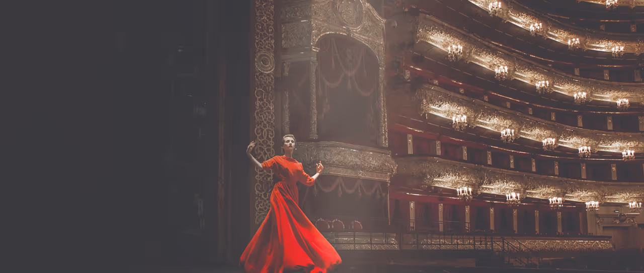 Bailarina em vestido vermelho dançando em um palco de teatro luxuoso com várias varandas e iluminação elaborada.