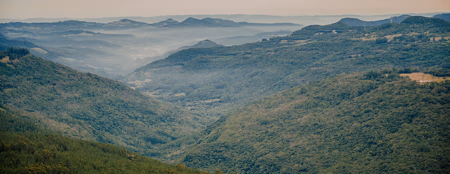Vista panorâmica de vales e colinas cobertos por densa vegetação verde com neblina leve e montanhas ao fundo.