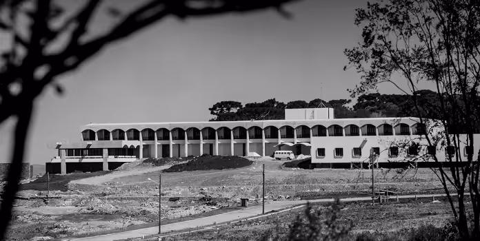 Prédio grande com colunas e arcos repetidos, em área aberta com árvores ao redor, em fotografia preto e branco.