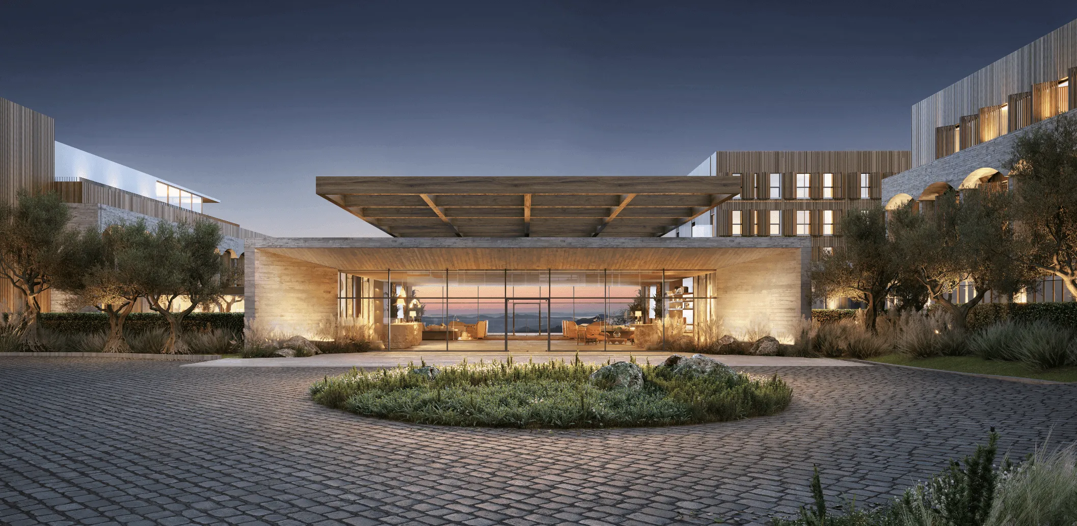 Entrada moderna de edifício com fachada de vidro iluminada ao entardecer e jardim circular com plantas e pedras.