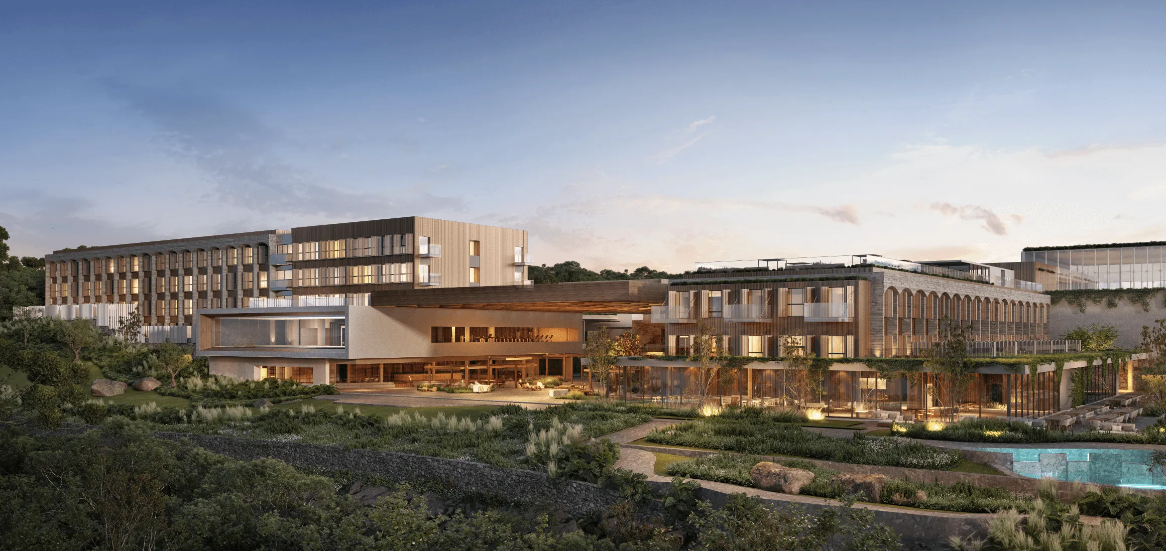 Vista externa de um complexo de edifícios modernos com iluminação interna, rodeado por jardins e uma piscina à direita ao entardecer.
