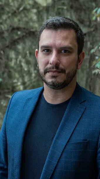 Homem de barba por fazer vestindo blazer azul e camiseta preta, posando ao ar livre com fundo de árvores desfocado.
