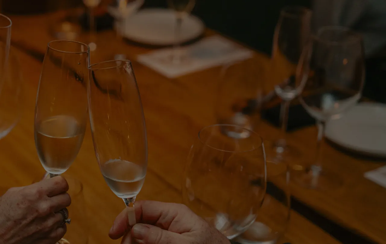 Duas pessoas brindando com taças de champanhe em uma mesa de madeira com outras taças e pratos ao fundo.