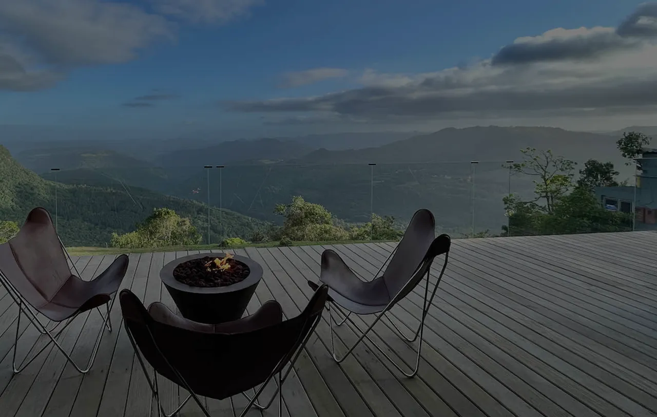 Deck de madeira com três cadeiras em volta de uma lareira ao ar livre com vista para montanhas verdes sob céu azul com nuvens.