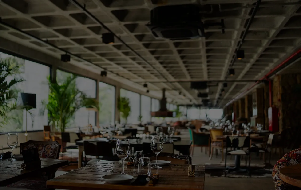 Restaurante espaçoso com mesas de madeira, cadeiras variadas, taças de vinho e plantas verdes ao longo das janelas grandes.