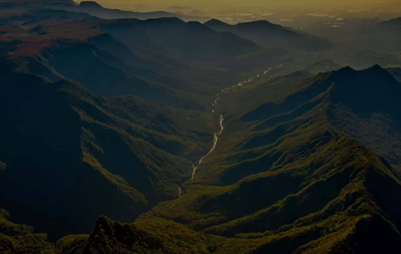 Vista aérea de uma cadeia montanhosa verde com um rio serpenteando pelo vale ao entardecer.