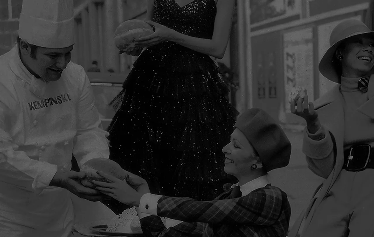 Chef sorridente entregando pão para uma mulher sentada usando boina, com outras duas mulheres ao redor segurando pães.