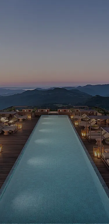 Piscina retangular iluminada ao entardecer com espreguiçadeiras e lanternas ao redor, com vista para montanhas ao fundo.