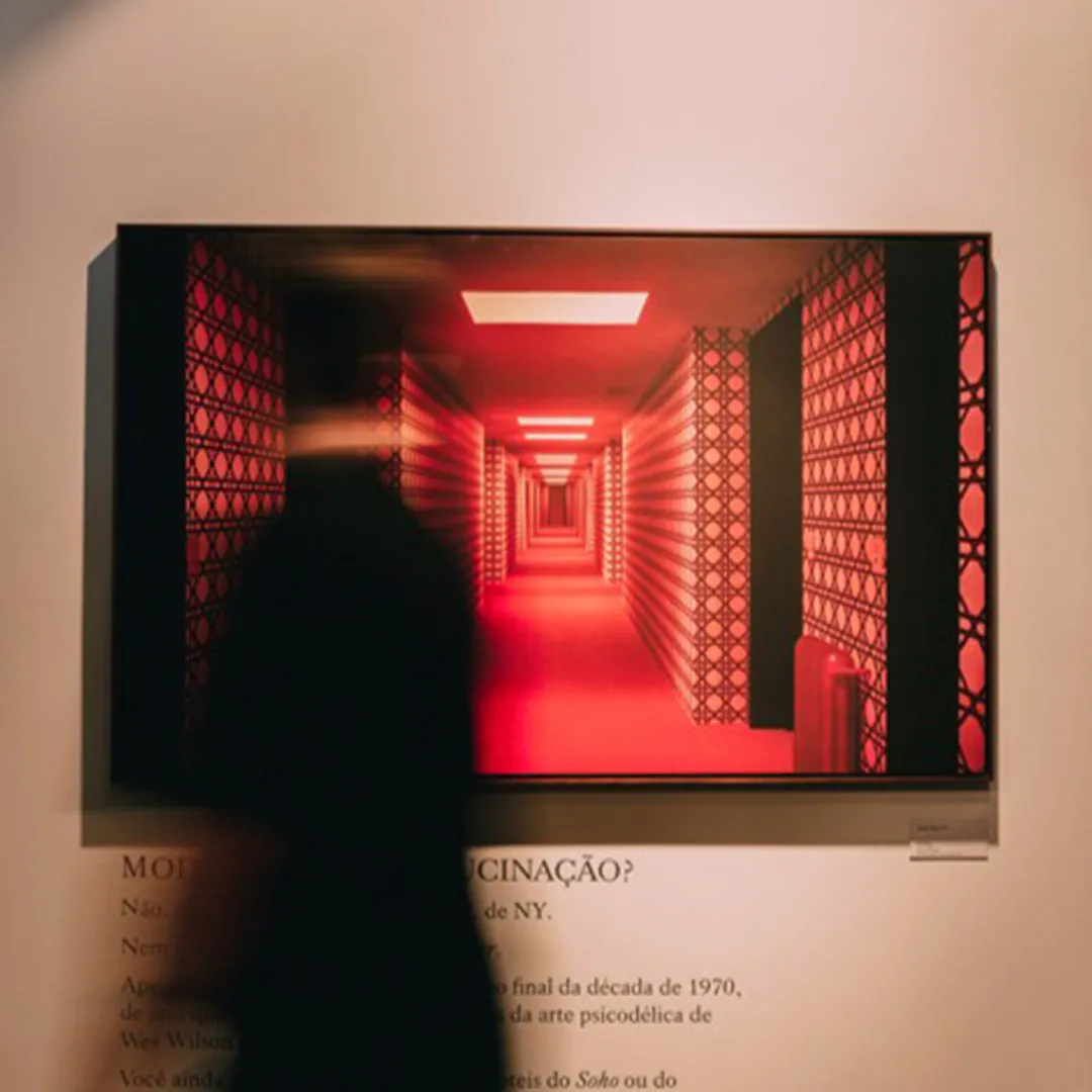 Pessoa desfocada passando em frente a uma obra de arte mostrando um corredor iluminado em tons vermelhos e paredes com padrão geométrico.
