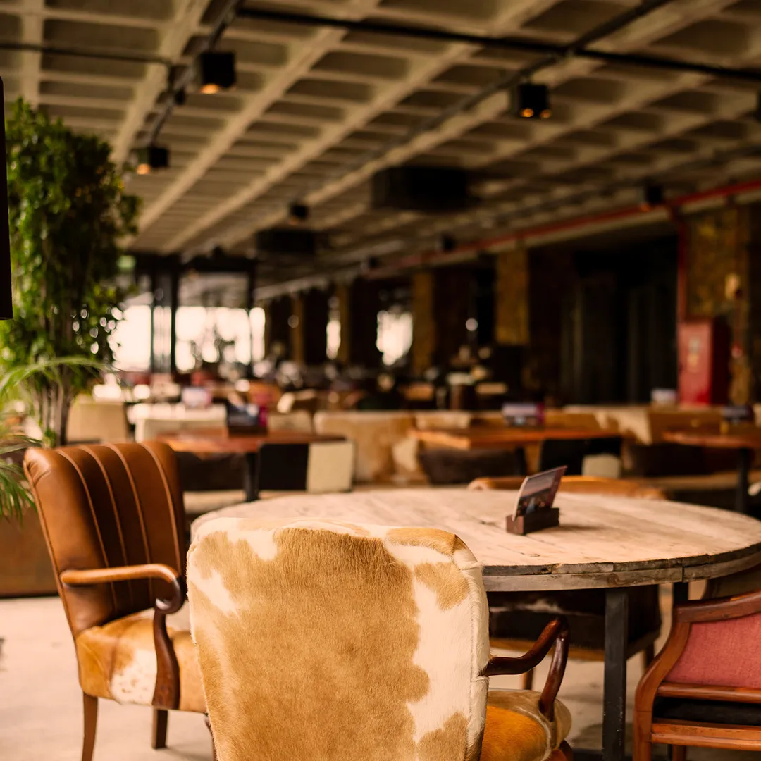 Interior de restaurante com cadeiras estofadas em couro e pelo, mesas de madeira e ambiente aconchegante e iluminado.