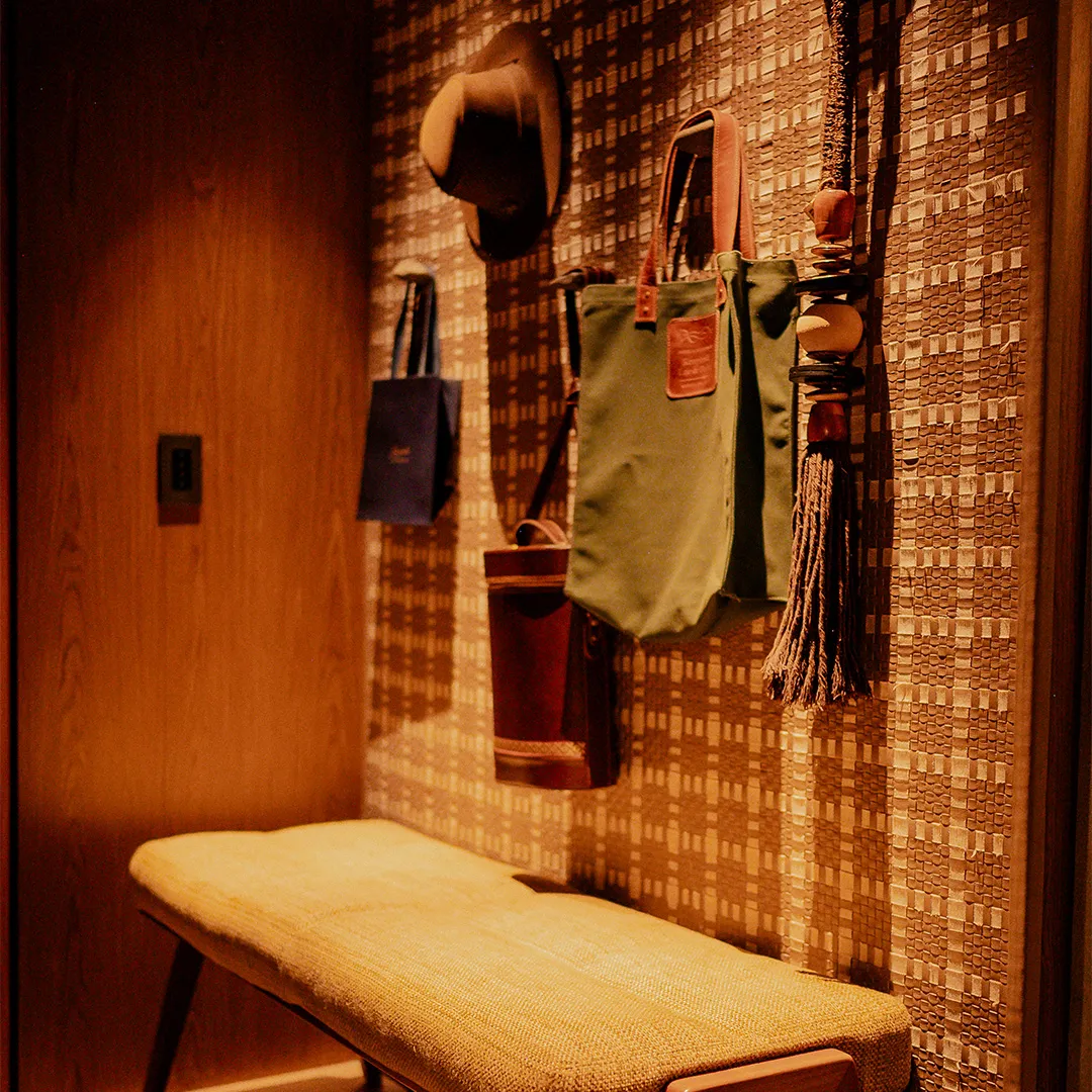 Banco estofado amarelo sob parede com papel de parede quadriculado e vários acessórios pendurados, incluindo chapéu e bolsas.