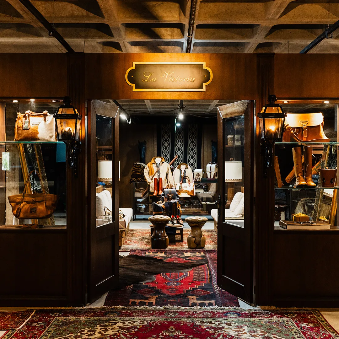 Entrada de loja La Victoria com portas de madeira abertas, exibindo bolsas, botas e roupas em um ambiente decorado com tapetes orientais.