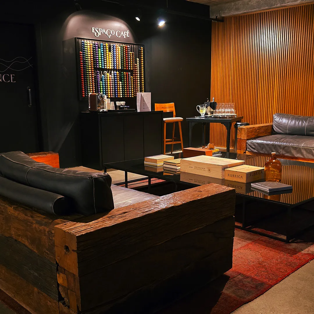Ambiente aconchegante de café com sofás de couro, mesa de centro preta com livros, parede preta com painel colorido de cápsulas de café e parede de madeira com venezianas.