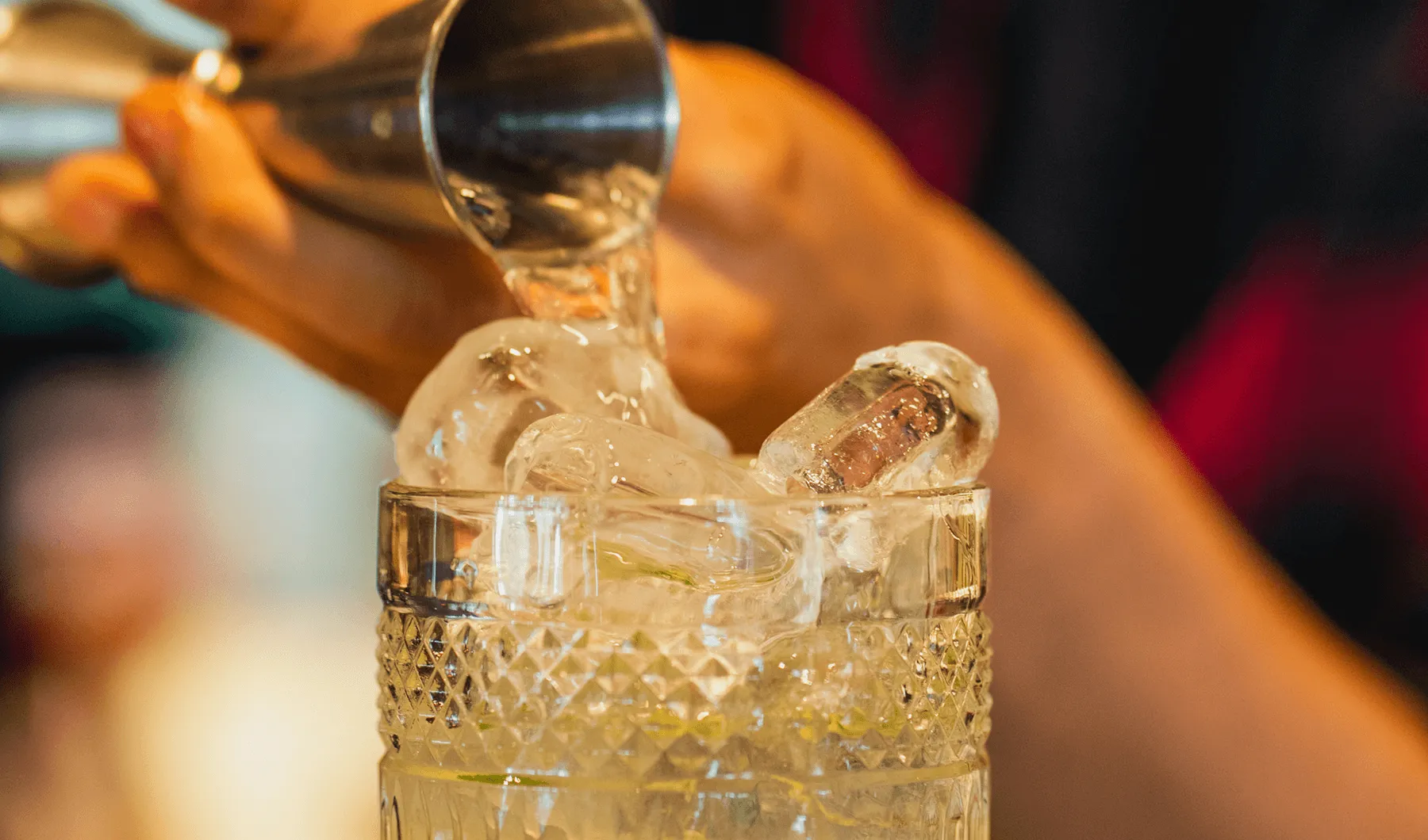Mão derramando líquido transparente sobre cubos de gelo em um copo de vidro texturizado.