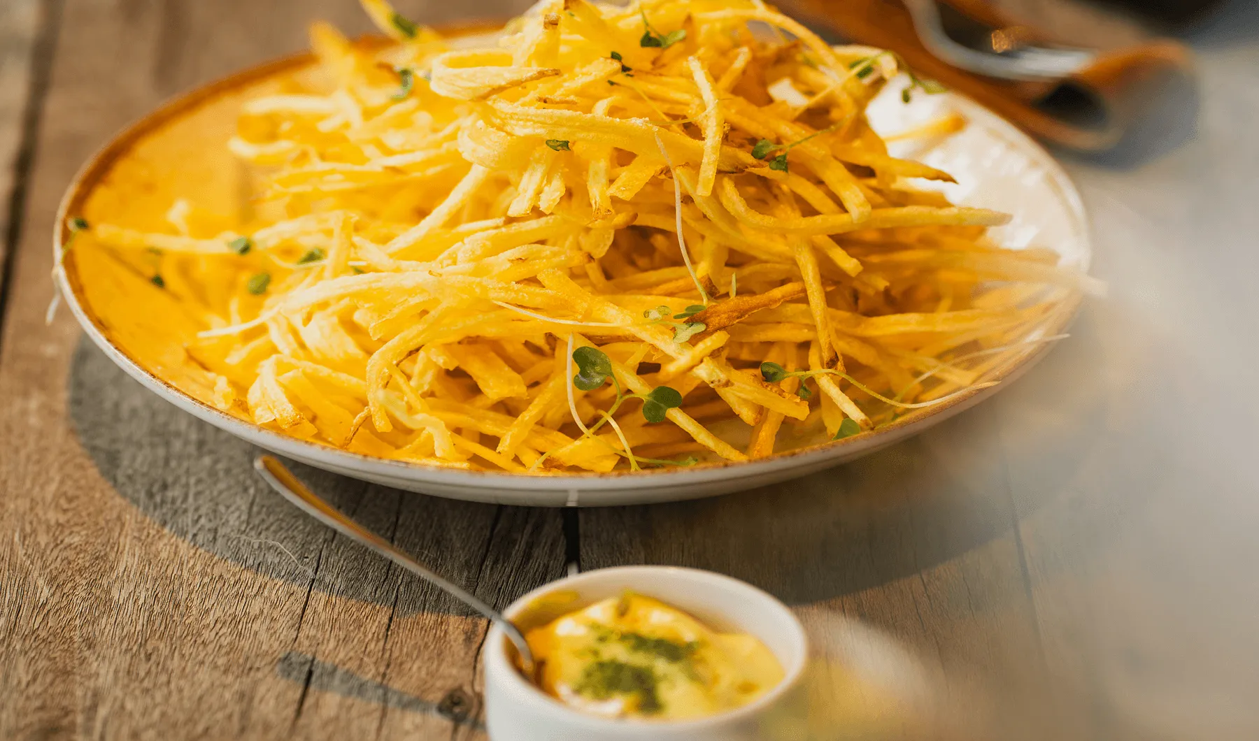 Tigela de batata palha dourada com ervas frescas servida com molho amarelo em recipiente branco.