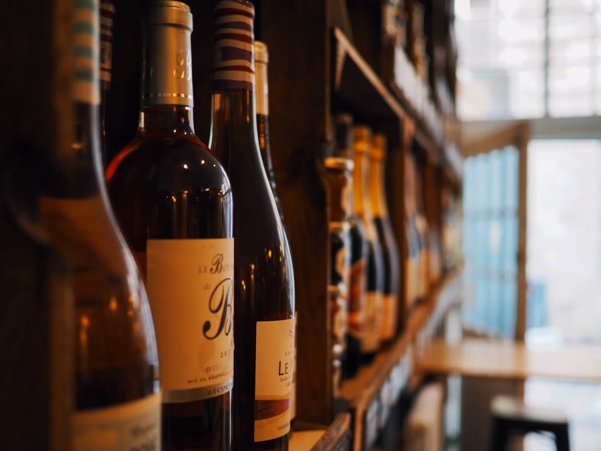Close-up of wine bottles on a wooden shelf in a dimly lit room with a window in the background.