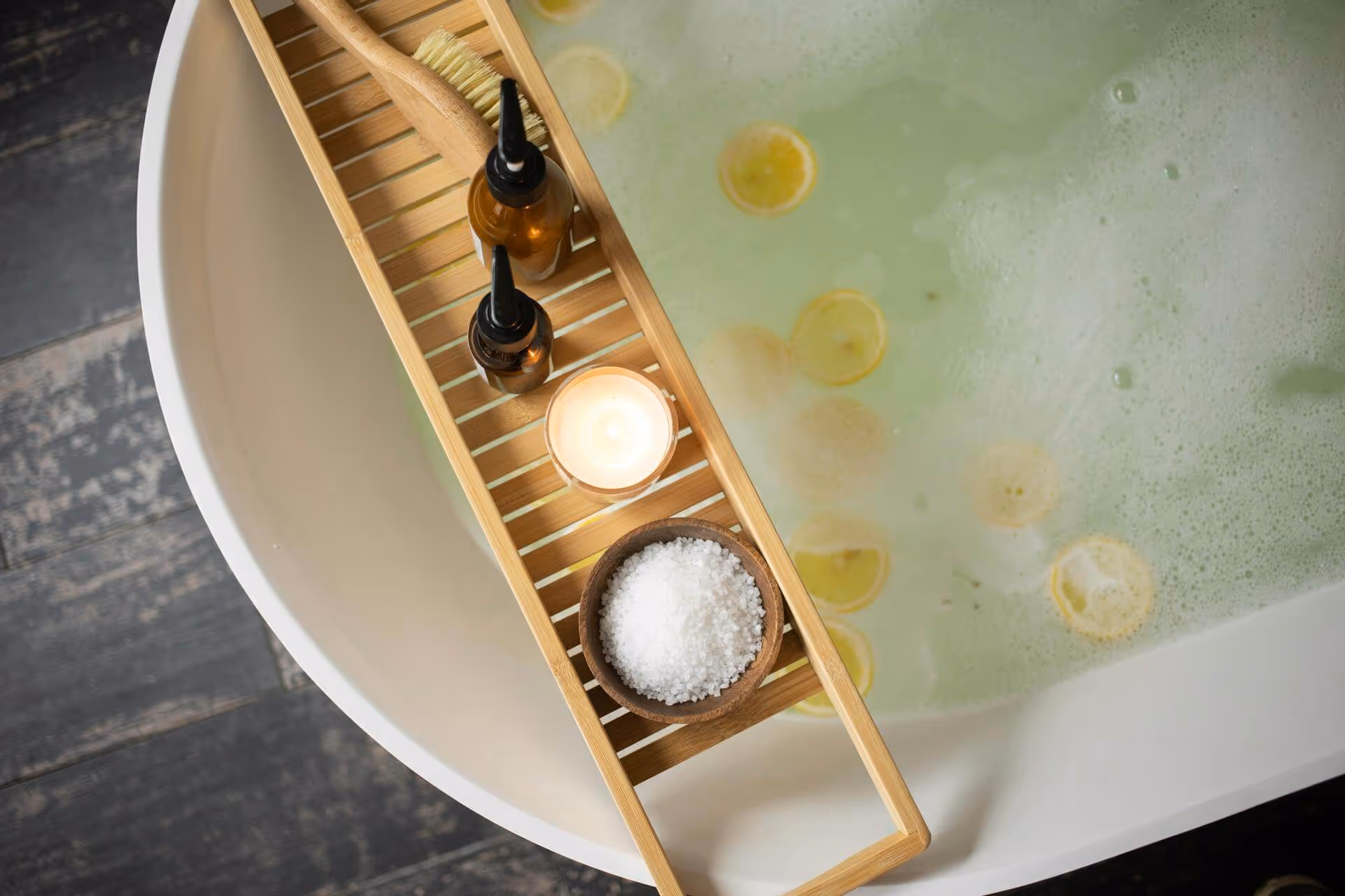 Wooden bath tray across a white bathtub filled with bubble bath and lemon slices, holding bath salt, a lit candle, two amber bottles, and a wooden brush.