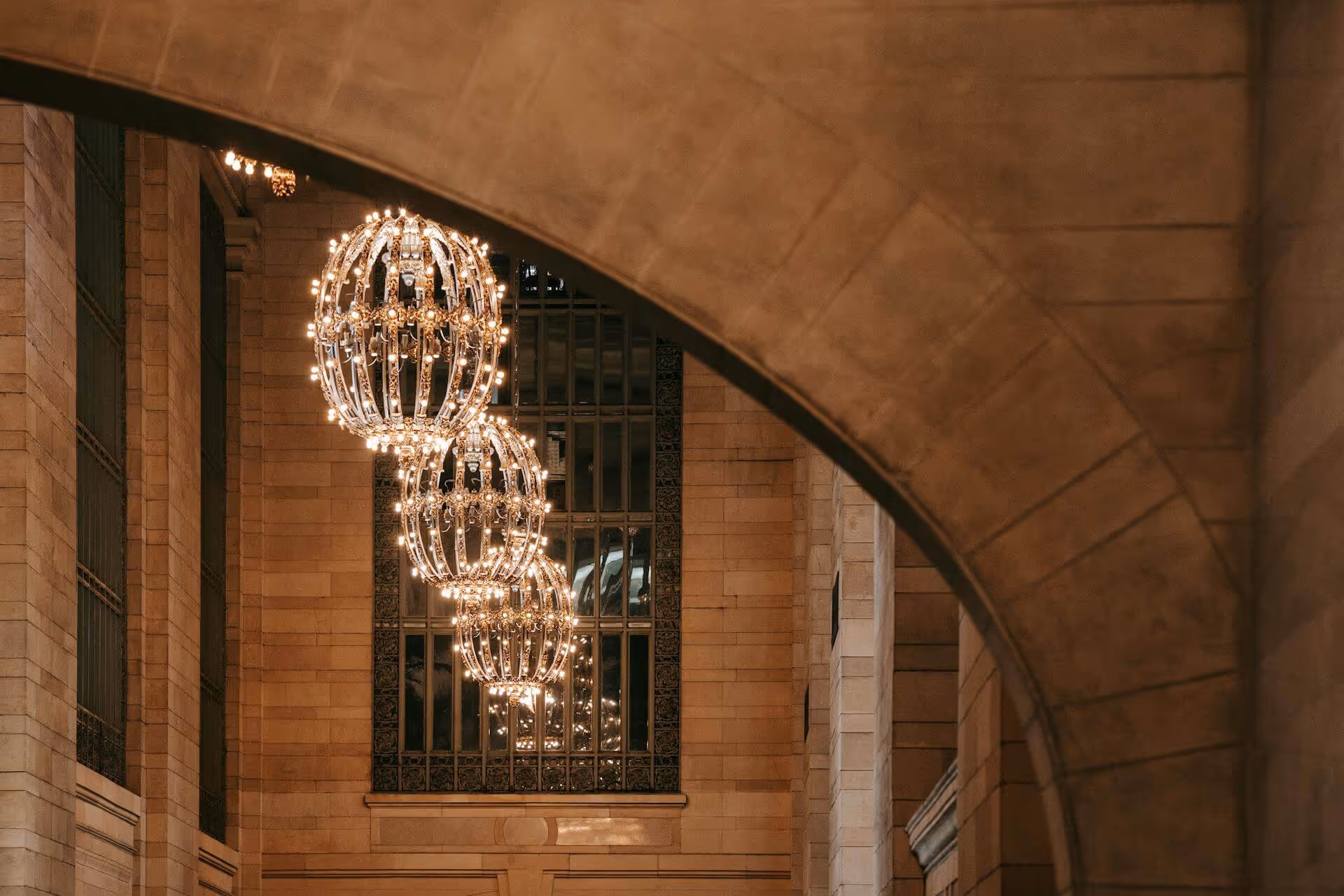 Three ornate spherical chandeliers hanging in a grand stone-arched hallway with large windows.