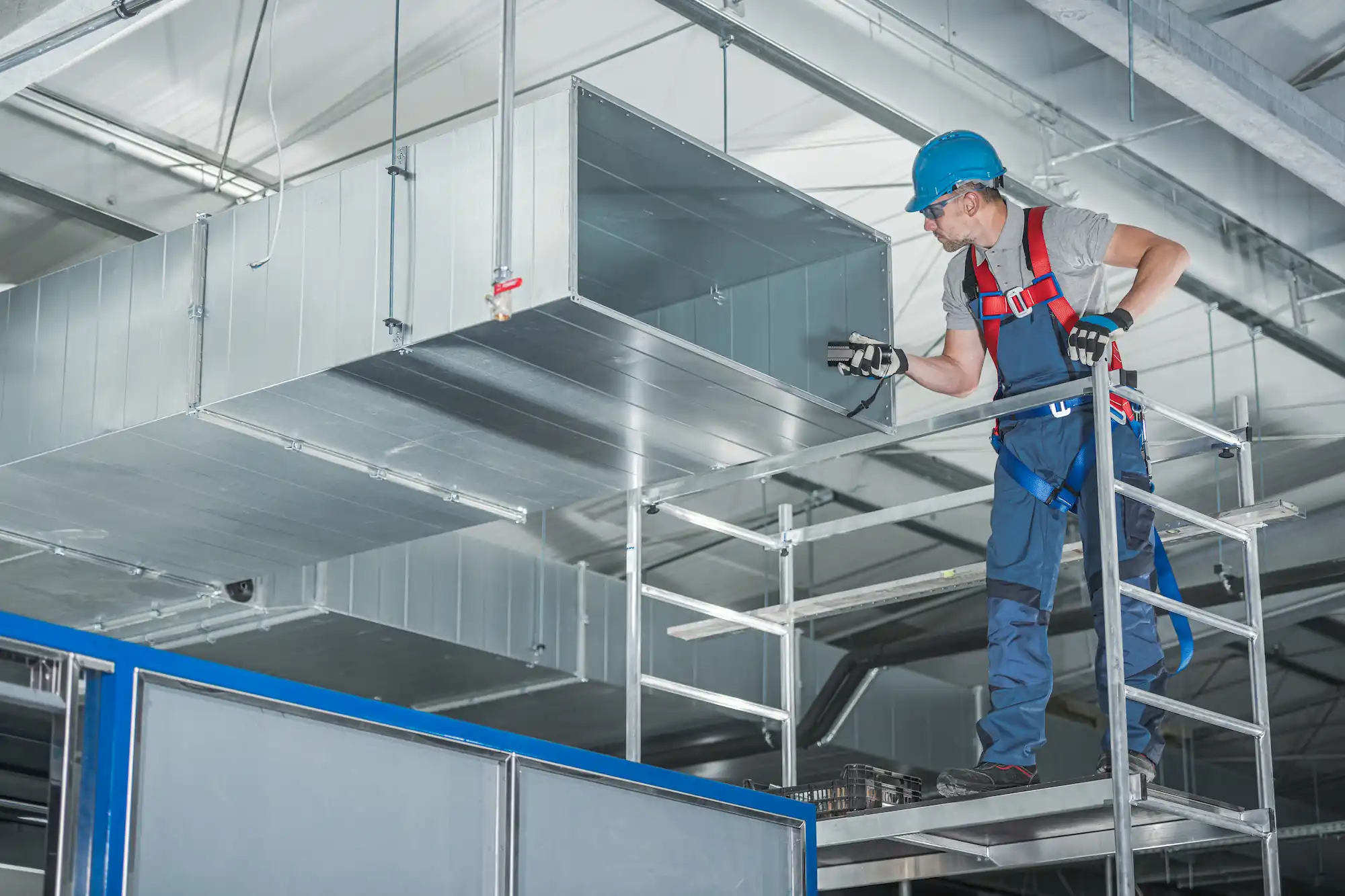 HVAC technician wearing a safety harness and helmet inspecting a large metal air duct inside an industrial building.