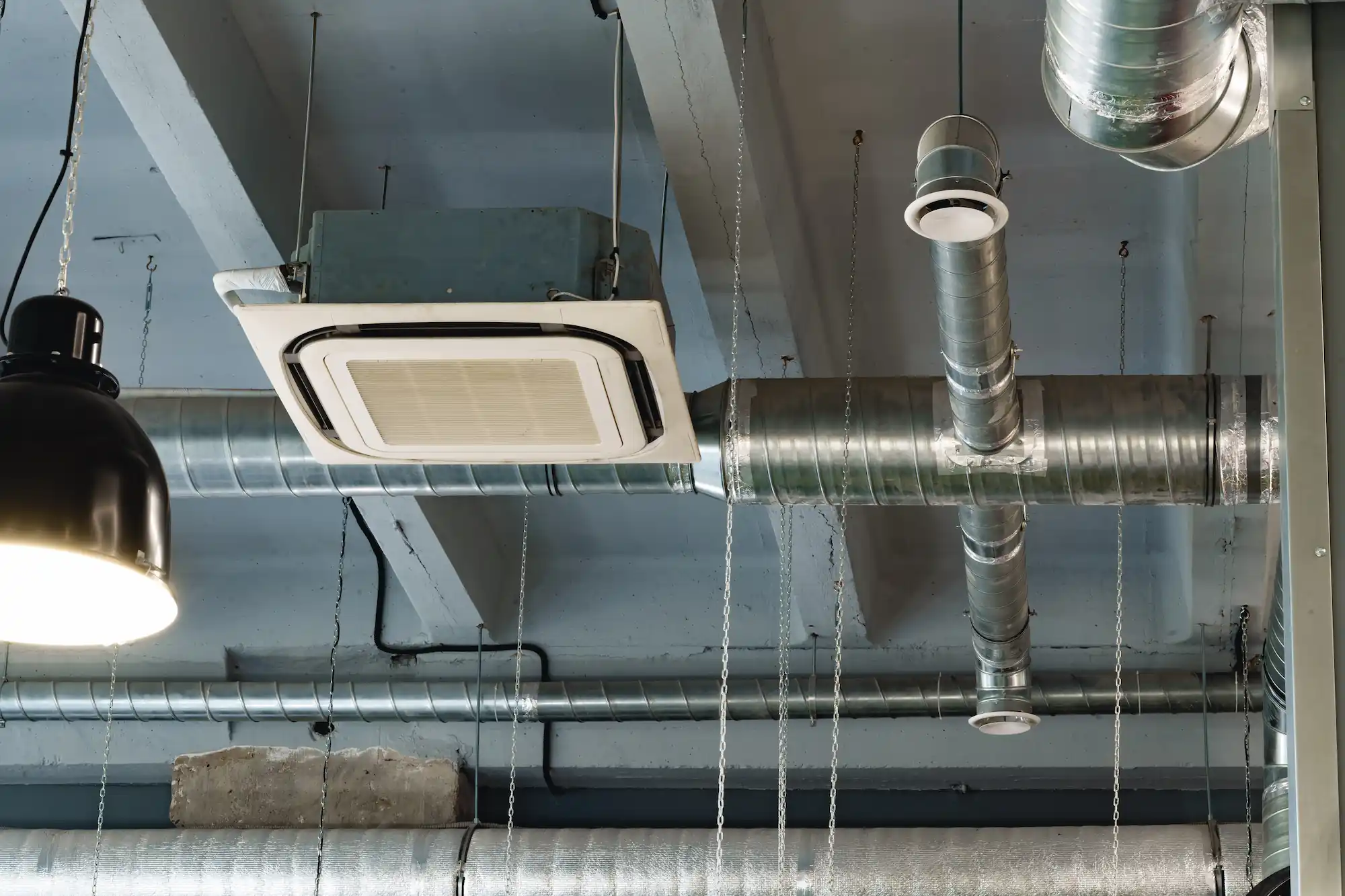 Silver ventilation pipes and an air conditioning unit mounted on a gray industrial ceiling with hanging chains and a black ceiling light.