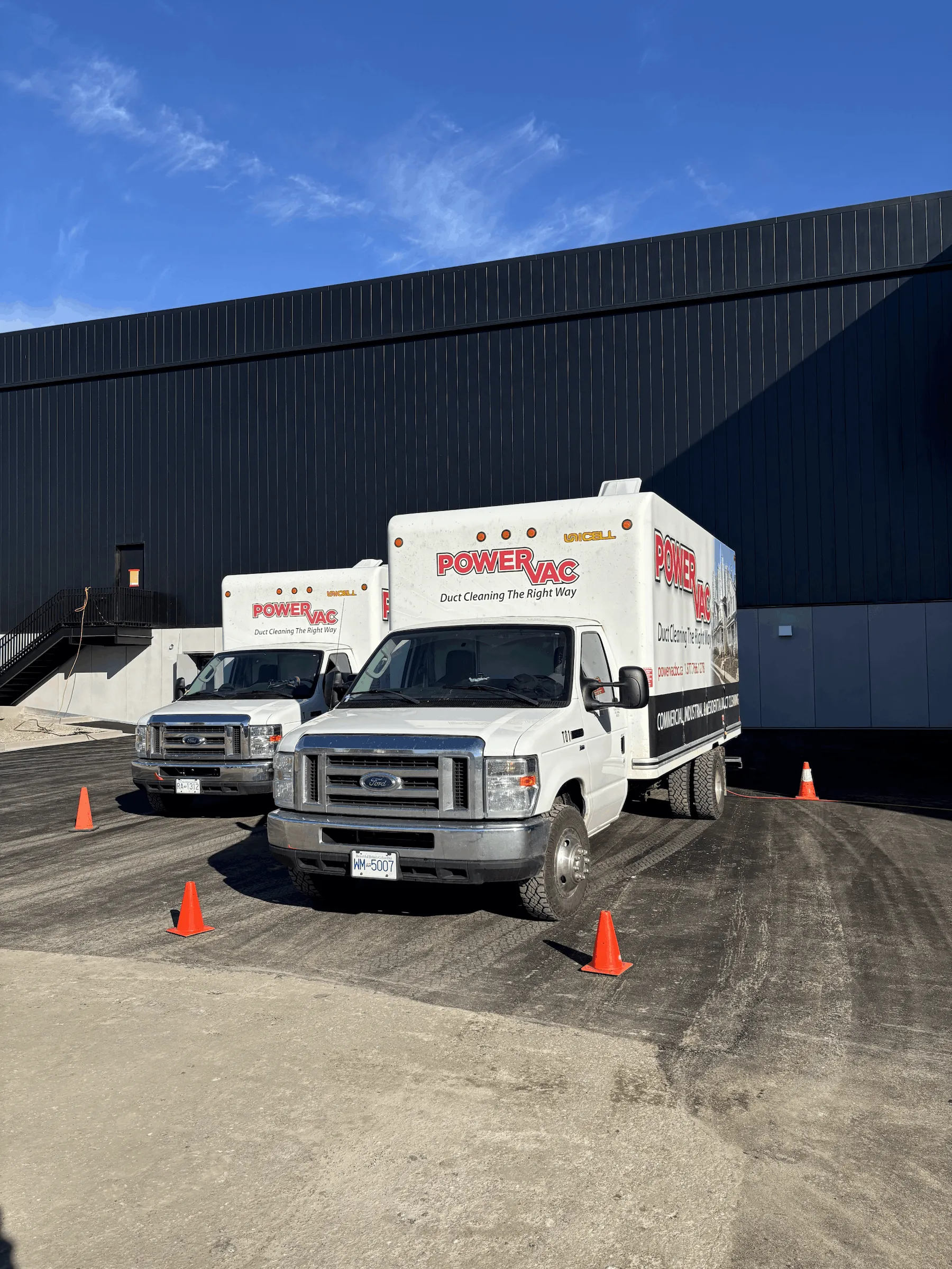 Two white PowerVac duct cleaning trucks parked outside a black industrial building with orange traffic cones around them.