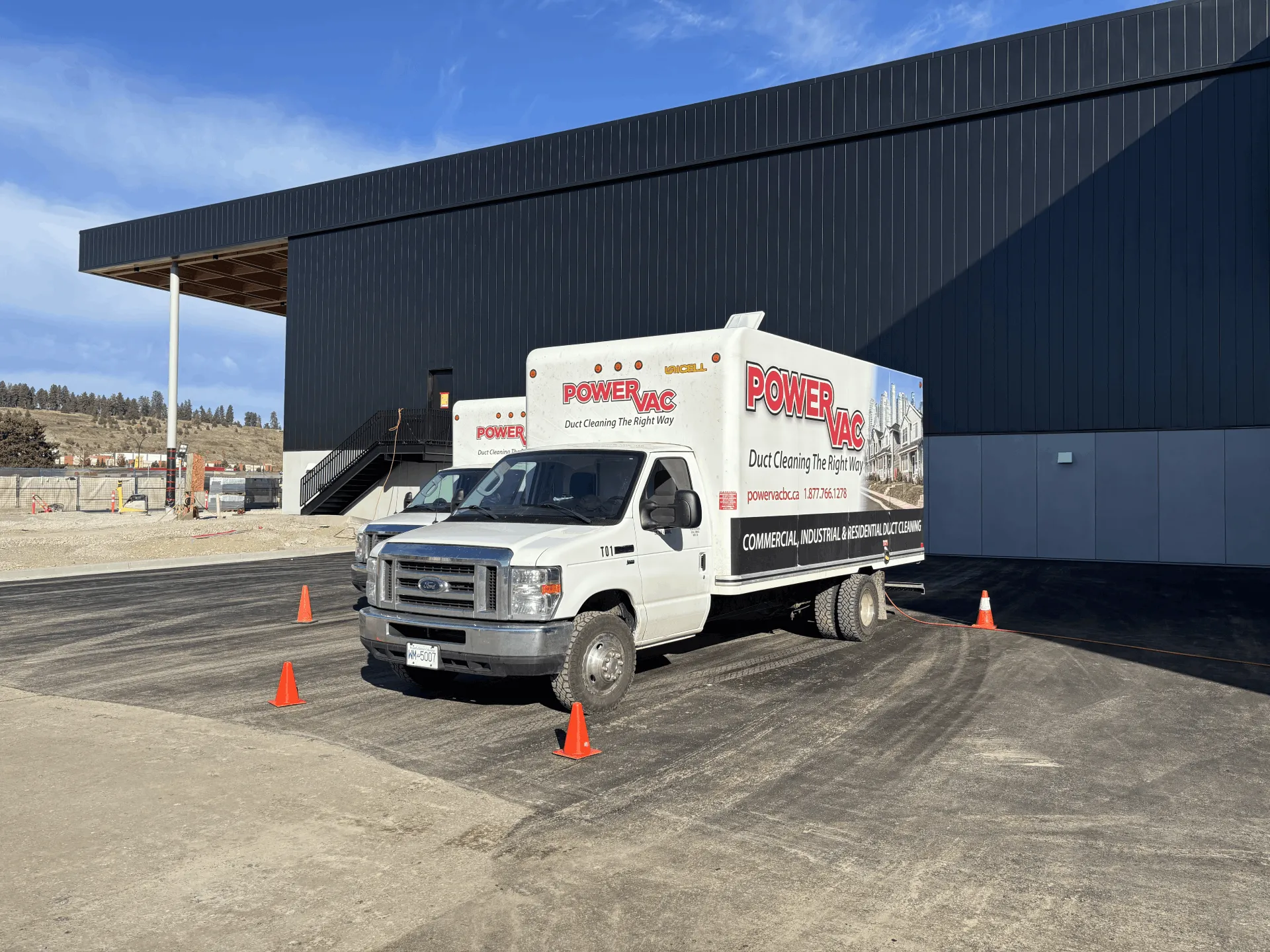 Two white PowerVac duct cleaning trucks parked outside a large dark building, surrounded by orange safety cones.