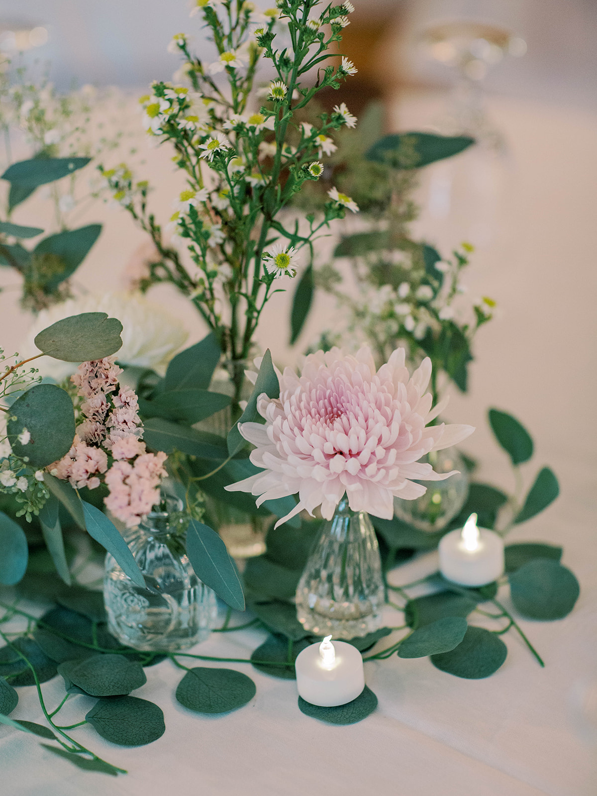 Table centerpiece with pink flowers, assorted greenery in glass vases, and two small white LED candles on a white tablecloth.