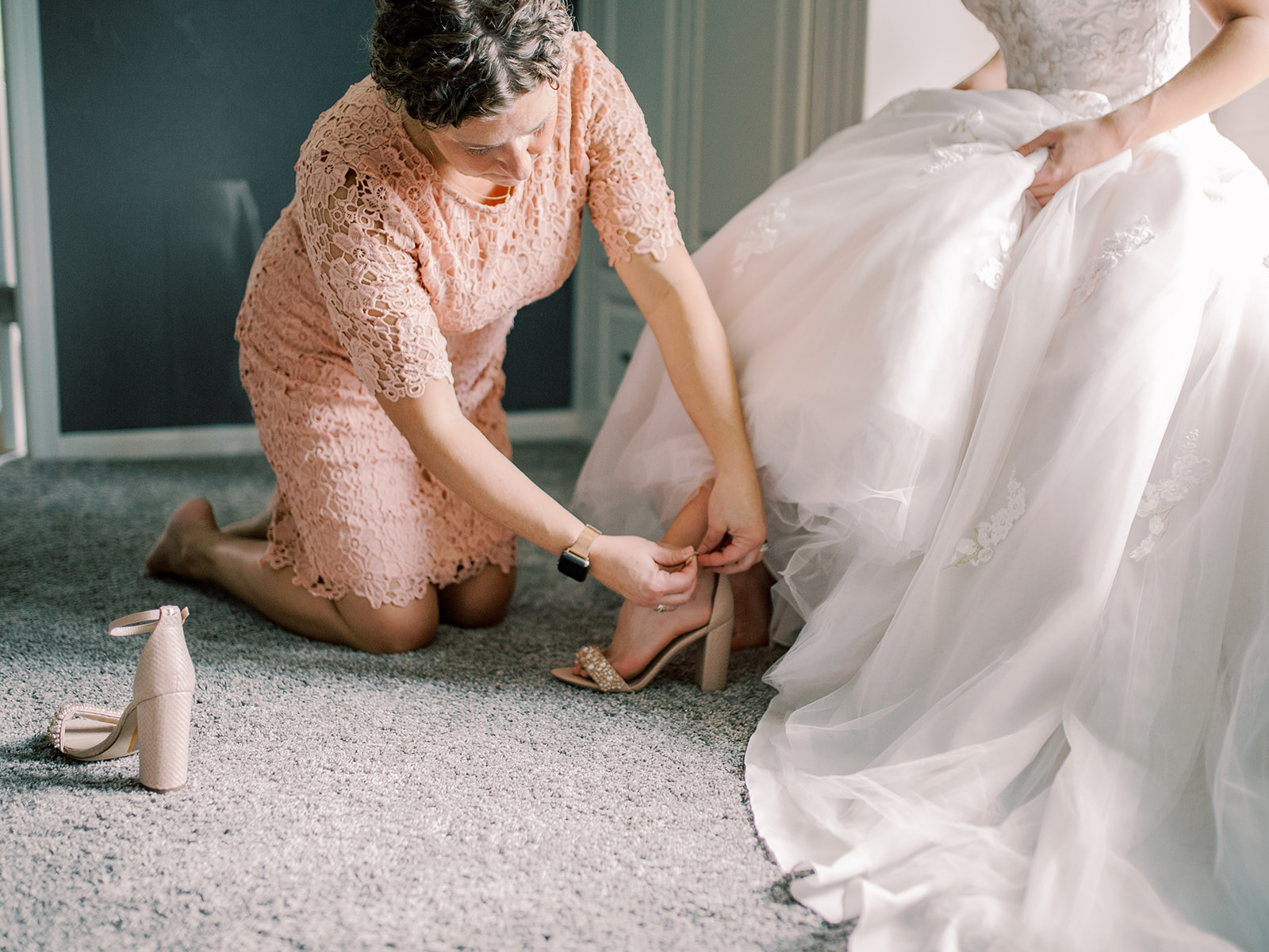 Woman in a peach lace dress kneeling on carpet, fastening a high-heeled shoe on a bride wearing a white wedding gown.