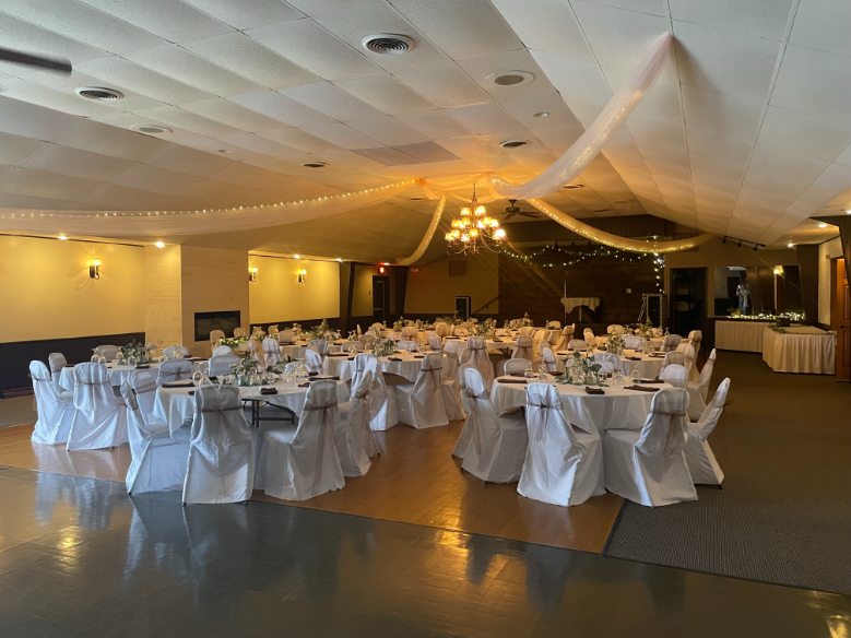 Banquet hall with round tables and white-covered chairs arranged for an event, decorated with draped fabric and string lights on the ceiling.