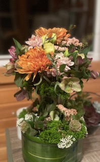 Floral arrangement in a green vase featuring orange chrysanthemums, pink and white flowers, and green foliage on a wooden surface.