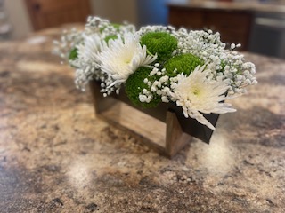 Small wooden holder with white chrysanthemums, green button flowers, and baby's breath on a brown granite countertop.