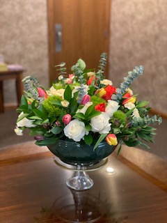 Elegant floral arrangement with white, red, and yellow flowers and green foliage in a glass pedestal vase on a wooden table.