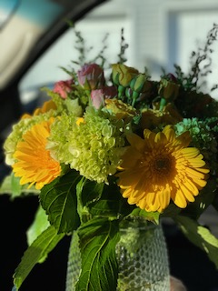 Close-up of a bouquet with yellow gerbera daisies, green flowers, and pink buds in a textured glass vase.