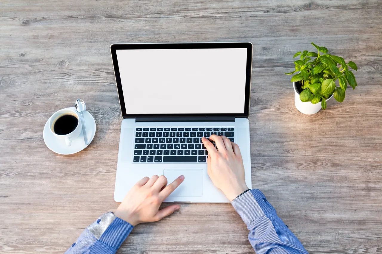 Top view of a person working on a laptop with coffee and a plant on a wooden desk.