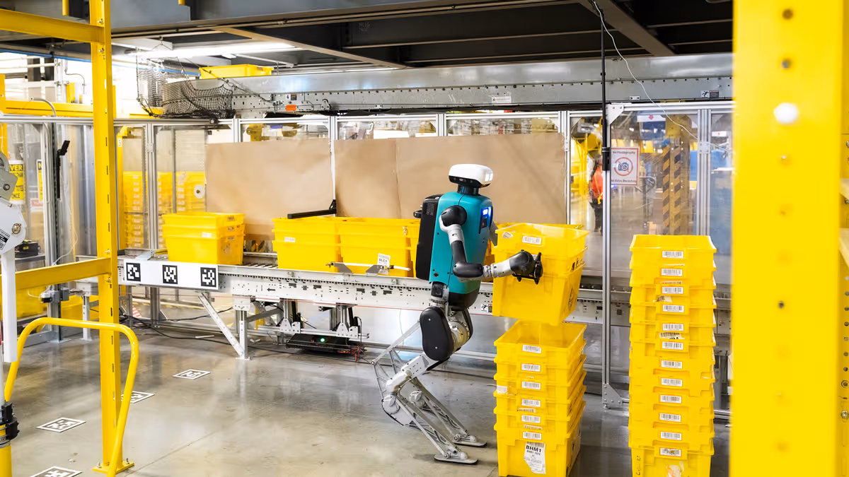 Humanoid robot with white head and teal torso stacking yellow bins in a warehouse environment.