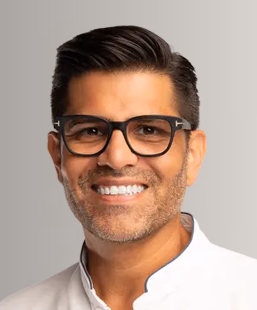Smiling man with short dark hair, wearing black glasses and a white collared shirt against a neutral background.