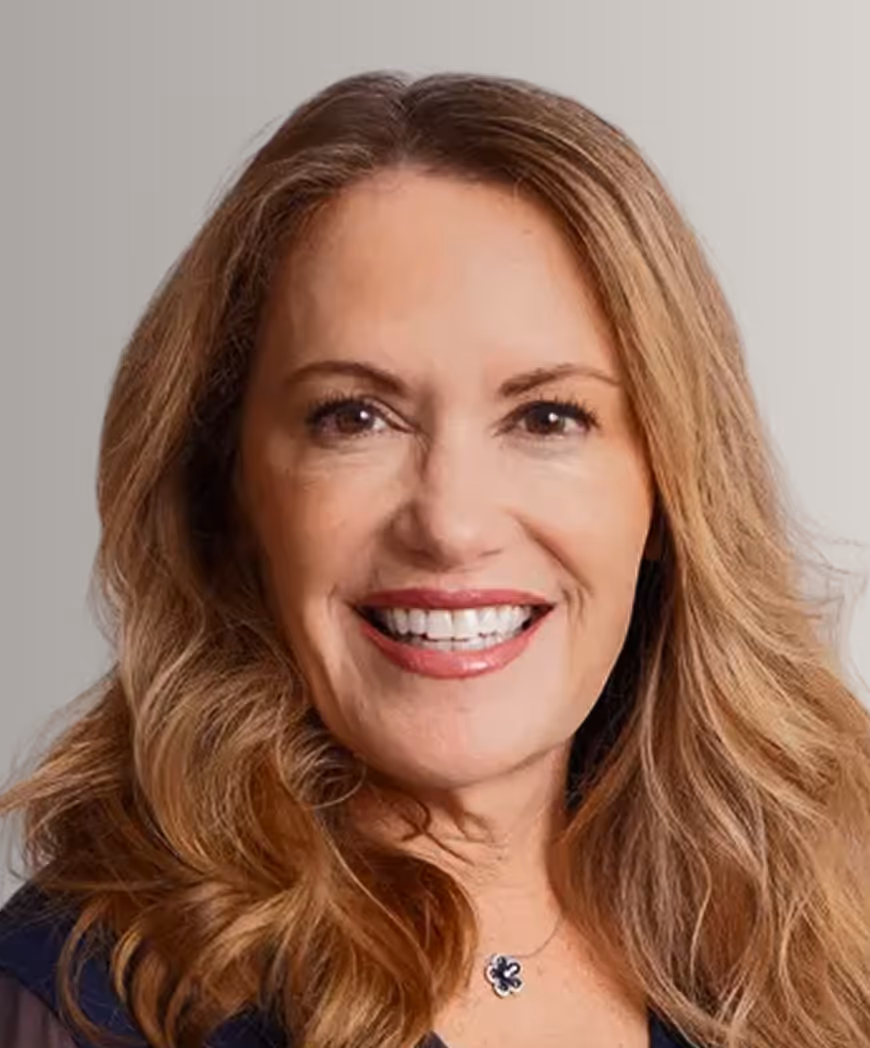 Smiling woman with long wavy blonde hair and a dark top, wearing a flower-shaped necklace.