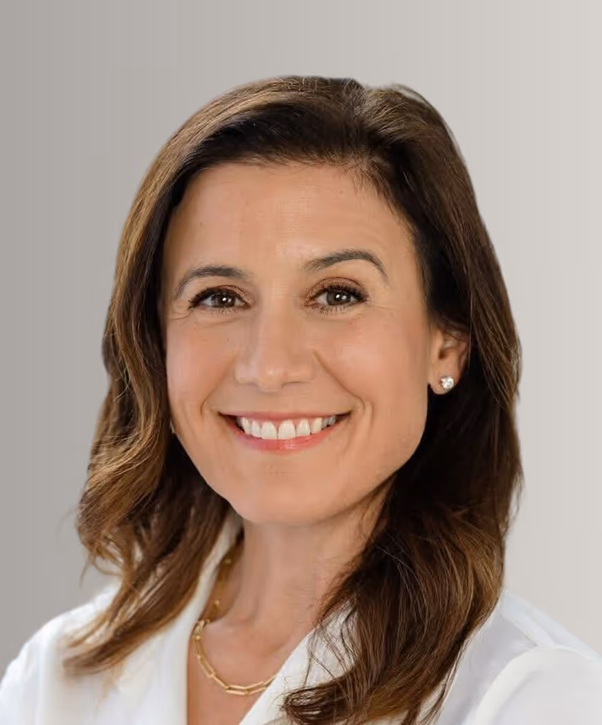 Smiling woman with medium-length brown hair wearing a white top and gold necklace against a light gray background.