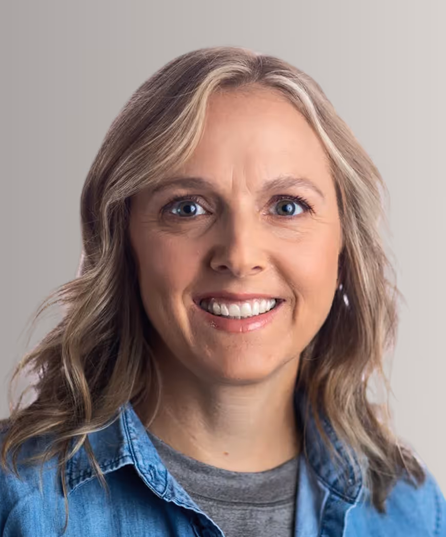 Smiling woman with shoulder-length blonde hair wearing a blue denim shirt over a gray top against a plain background.