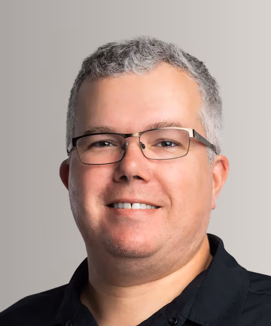 Smiling man with short gray hair and glasses wearing a black shirt against a plain light background.
