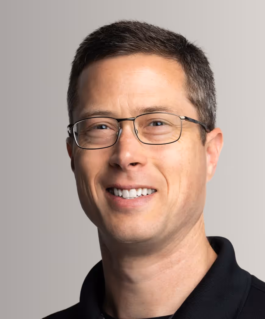 Smiling man with short brown hair and glasses wearing a black collared shirt against a plain background.