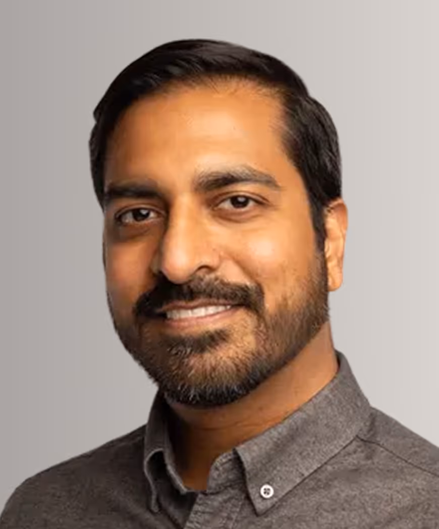 Smiling man with beard wearing a gray button-up shirt against a light gray background.