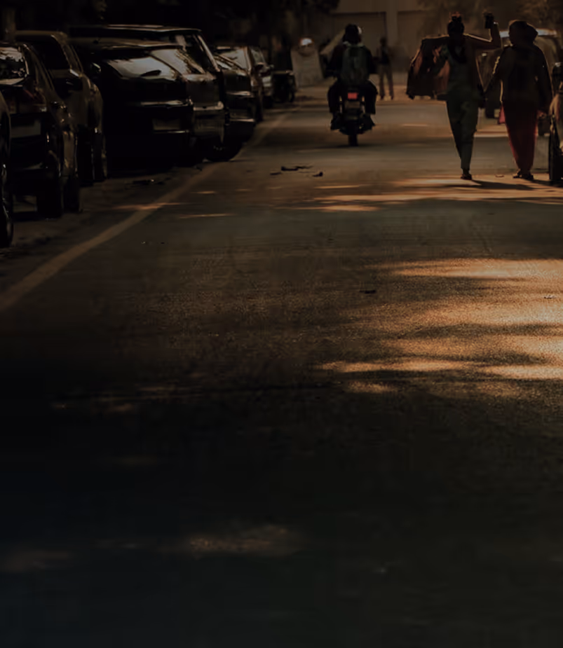 A dimly lit street with parked cars on the left, a motorcyclist riding away, and two women walking hand in hand on the right side.