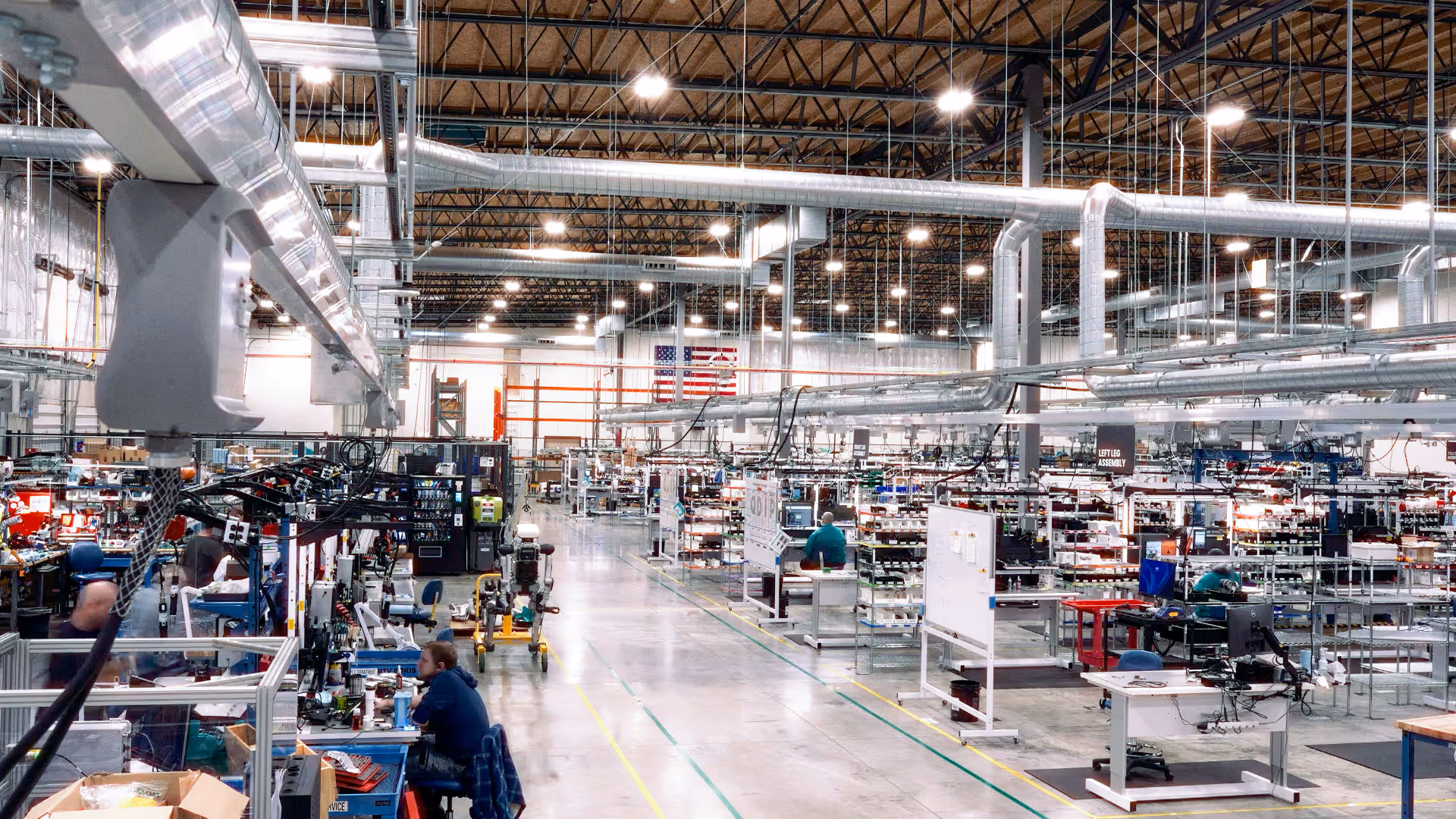Spacious, brightly lit manufacturing factory interior with workers at stations and overhead ductwork.