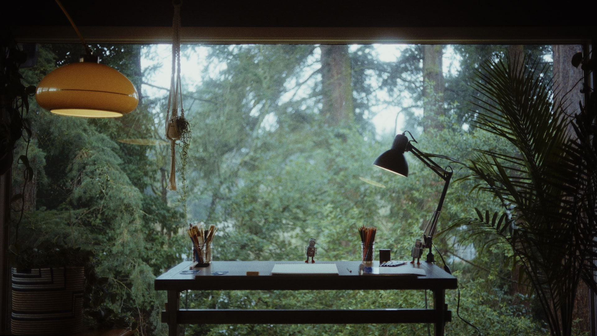 Minimalist desk with writing utensils, a lamp, and small robot figures positioned in front of a large window overlooking dense green forest.