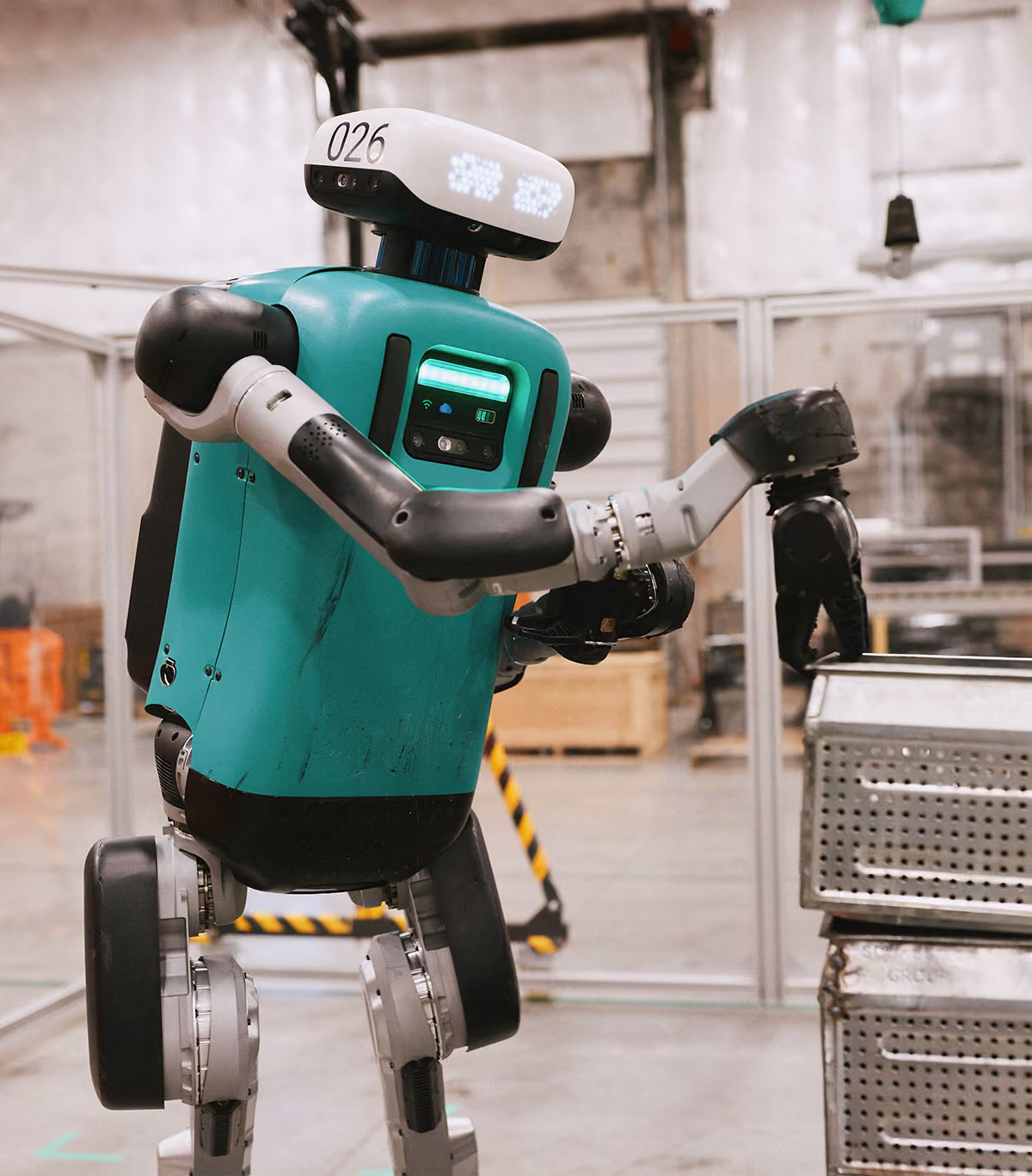A teal and black humanoid robot working inside an industrial facility, reaching toward a metal bin.