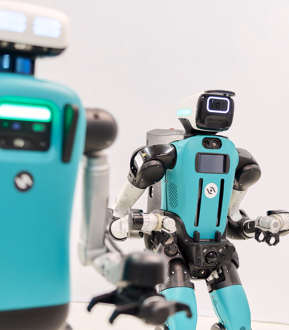 Two humanoid robots with teal and black bodies facing forward against a plain light background, one robot is in clear focus showing detailed arms and head with a screen-like face.